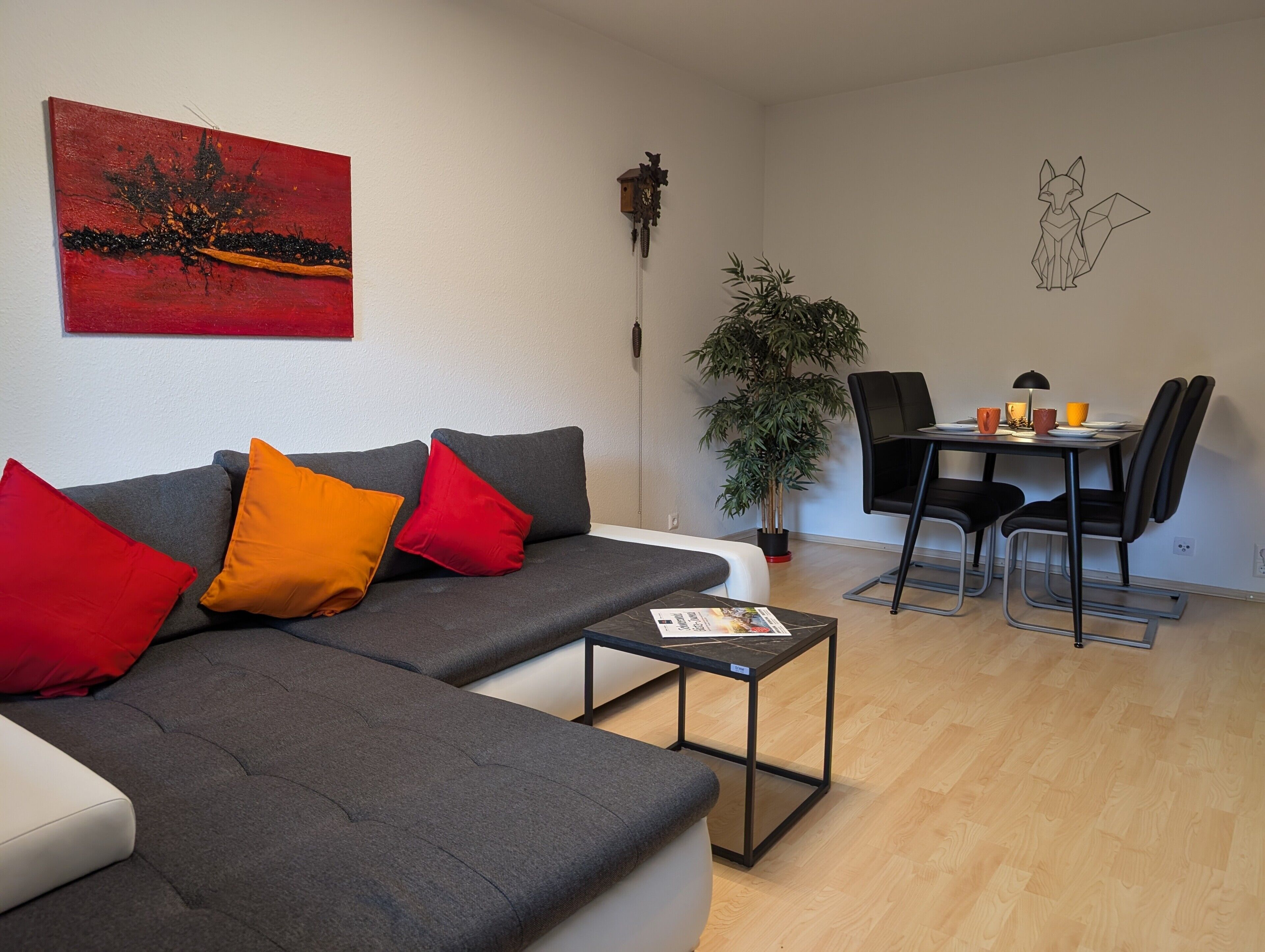 A modern living room with a grey sofa, red and orange cushions, a small table, a dining area with a black chair, and a potted plant.