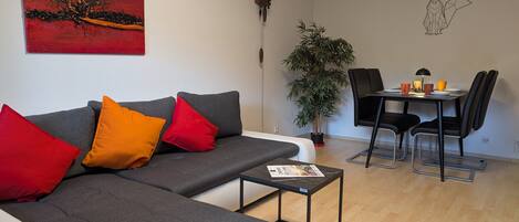 A modern living room with a grey sofa, red and orange cushions, a small table, a dining area with a black chair, and a potted plant.