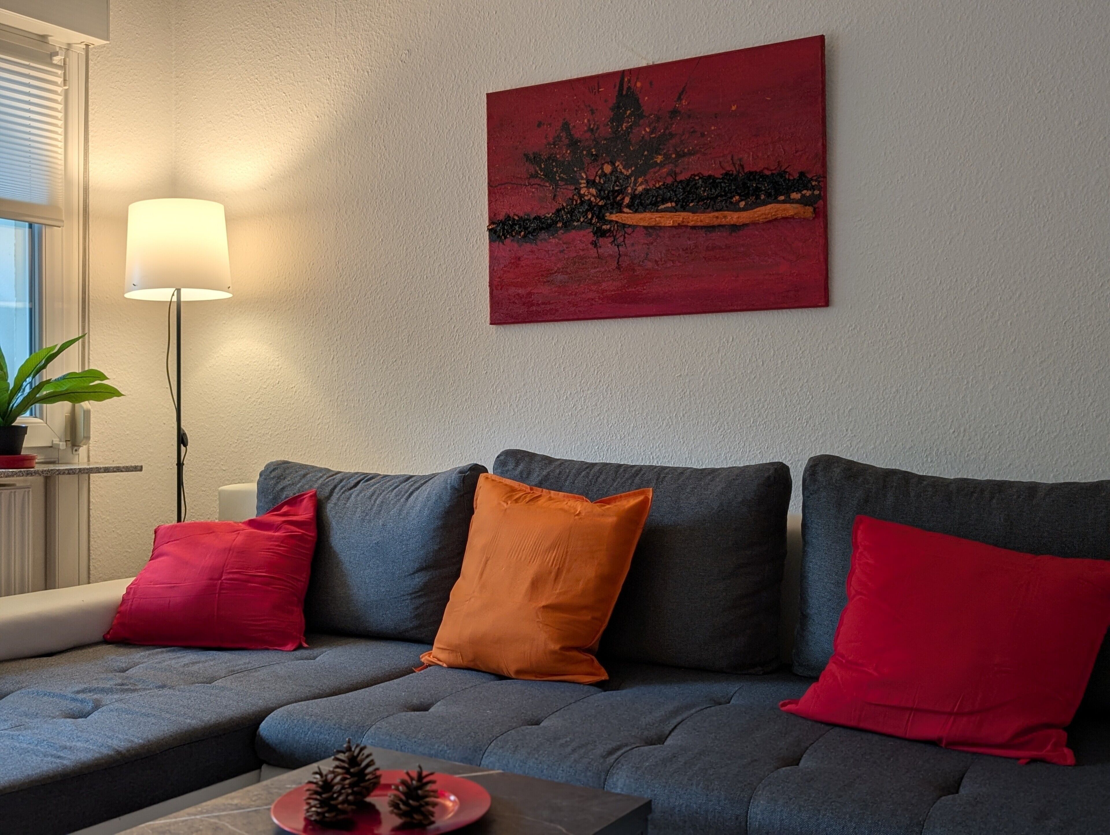A grey sofa with red and orange cushions, a painting on the wall, a floor lamp, and a small table with pine cones.