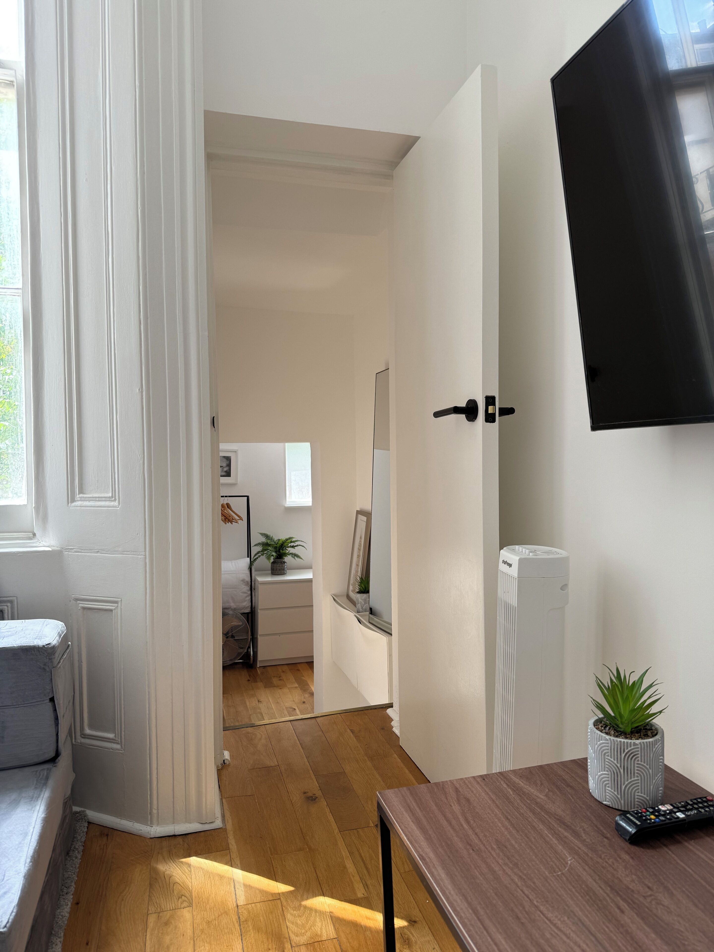 A room with a wooden floor, a white door, a bed, a bedside table, a potted plant, a TV mounted on the wall, and a radiator.