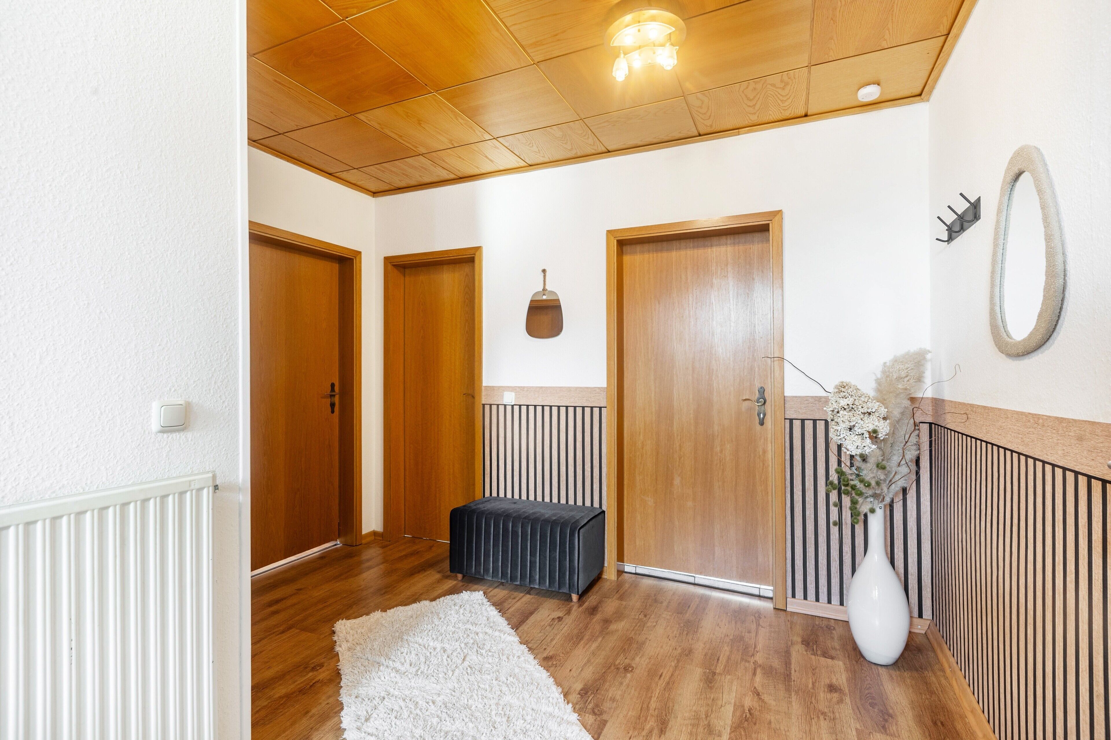 A hallway with wooden doors, a radiator, a white rug, a black bench, and a vase with flowers.