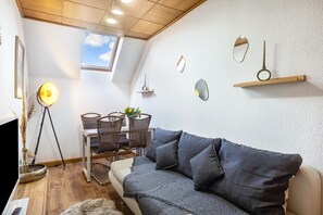 A cozy room with a sofa, a dining table with chairs, a lamp, and a small shelf with decorative items.