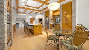 A modern kitchen with wooden cabinets, a central island, and a dining area with wicker chairs and a glass table. - The Manor-Indoor Pool-Speak Easy-Game Room-Sauna (Bentonville)