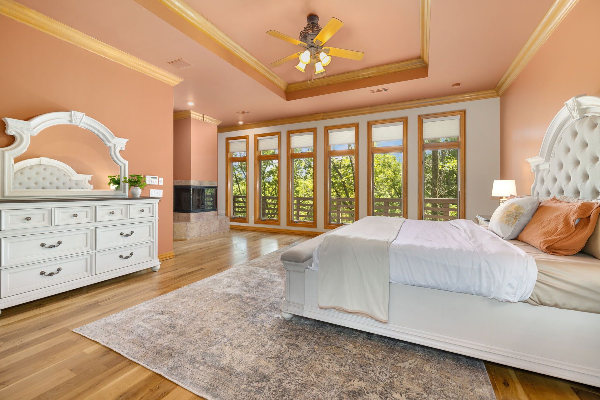 Ein Schlafzimmer mit einem großen Bett, einem Schminktisch, einem Deckenventilator und Blick auf Bäume durch große Fenster.