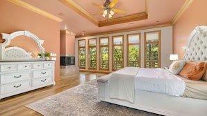 A bedroom with a large bed, a dresser, a ceiling fan, and a view of trees through large windows. - The Manor-Indoor Pool-Speak Easy-Game Room-Sauna (Bentonville)