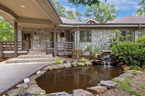 A stone house with a pond, wooden deck, and a covered porch. - The Manor-Indoor Pool-Speak Easy-Game Room-Sauna (Bentonville)