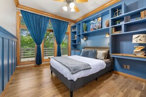 A bedroom with a bed, blue curtains, a ceiling fan, and a bookshelf with various items. - The Manor-Indoor Pool-Speak Easy-Game Room-Sauna (Bentonville)