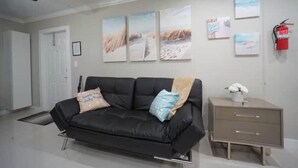 A living room with a black leather sofa, a small bedside table with a vase of flowers, and three framed pictures on the wall. - Modern Beach Studio 3 (Pompano Beach)