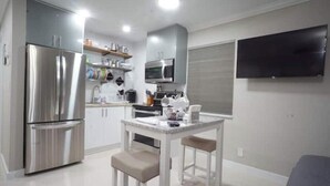 A modern kitchen with stainless steel appliances, a white dining table, and a flat-screen TV mounted on the wall. - Modern Beach Studio 3 (Pompano Beach)
