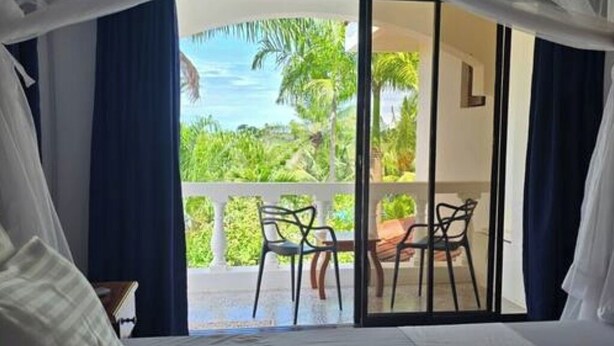 Ein Schlafzimmer mit einem großen Bett, einem Balkon mit Sitzgelegenheiten und Blick auf Grünflächen und Palmen.