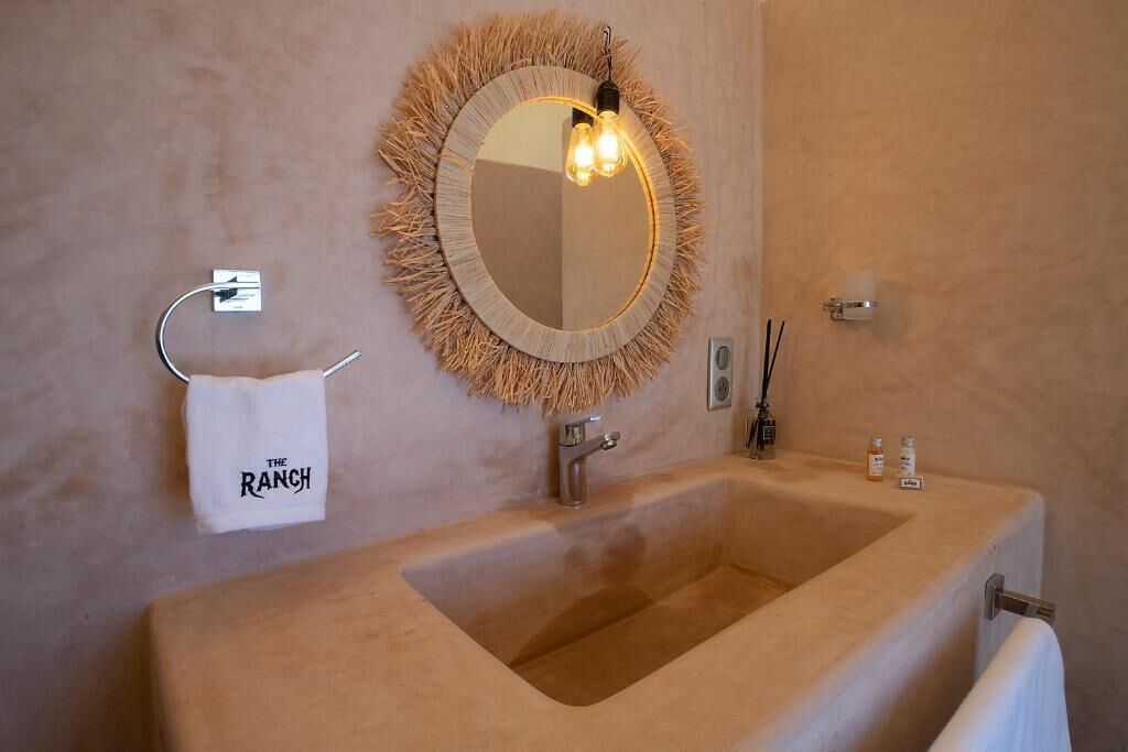 A bathroom with a large beige sink, a round mirror with a fringe, a towel rack with a towel labeled 'The Ranch', and a wall-mounted light fixture.