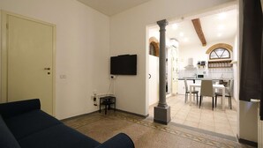 Una sala de estar con un sofá, un televisor montado en la pared y una cocina visible a través de un vano abierto.