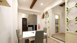 Un comedor con una mesa blanca y cuatro sillas, un extintor en la pared, y vista a una cocina a través de una puerta abierta.