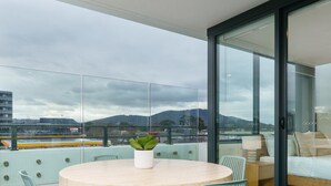En balkon med et rundt bord og stole, en potteplante og udsigt over et bybillede med bjerge i baggrunden.