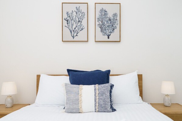 Ein ordentlich bezogenes Bett mit weißer Bettwäsche, zwei dekorative Kissen und ein blaues Akzentkissen. An der Wand über dem Bett befinden sich zwei gerahmte Pflanzenmotive.