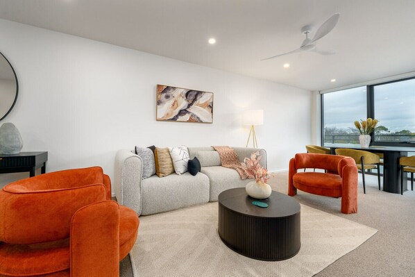 Ein modernes Wohnzimmer mit einem grauen Sofa, orangefarbenen Sesseln, einem schwarzen Couchtisch und einem großen Fenster mit Blick ins Freie.