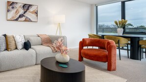 Ein modernes Wohnzimmer mit einem grauen Sofa, einem roten Sessel, einem runden Couchtisch und einem großen Fenster mit Blick ins Freie.