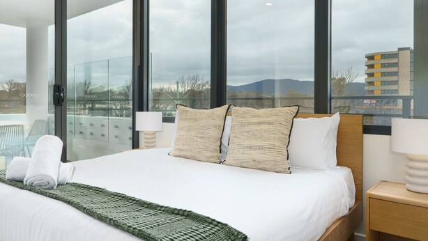 Ein modernes Hotelzimmer mit einem großen Bett, zwei Kissen, einer grünen Tagesdecke und Blick auf die Stadt durch große Fenster.