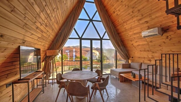 Une cabane en bois chaleureuse, dotée d’une grande fenêtre triangulaire, d’une table à manger ronde et d’un canapé.
