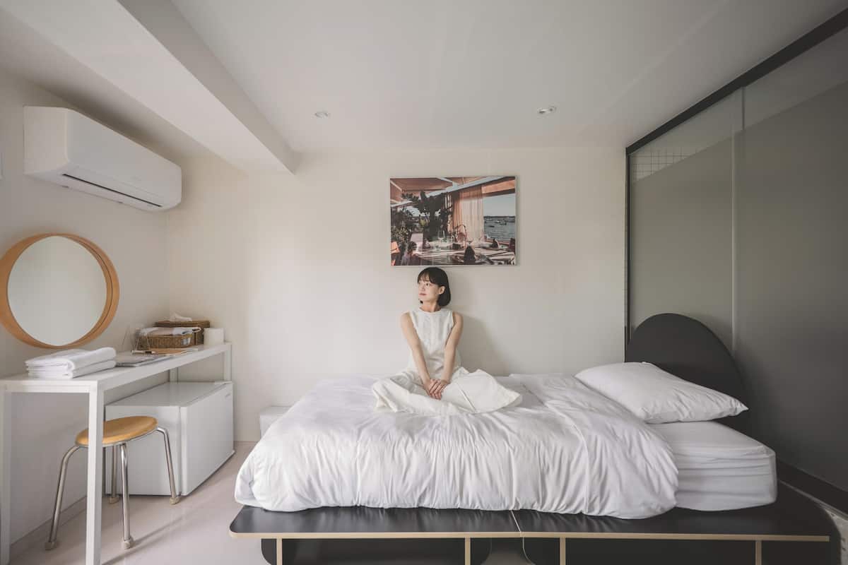 Un dormitorio moderno con cama, escritorio, silla, espejo redondo y aire acondicionado instalado en la pared.