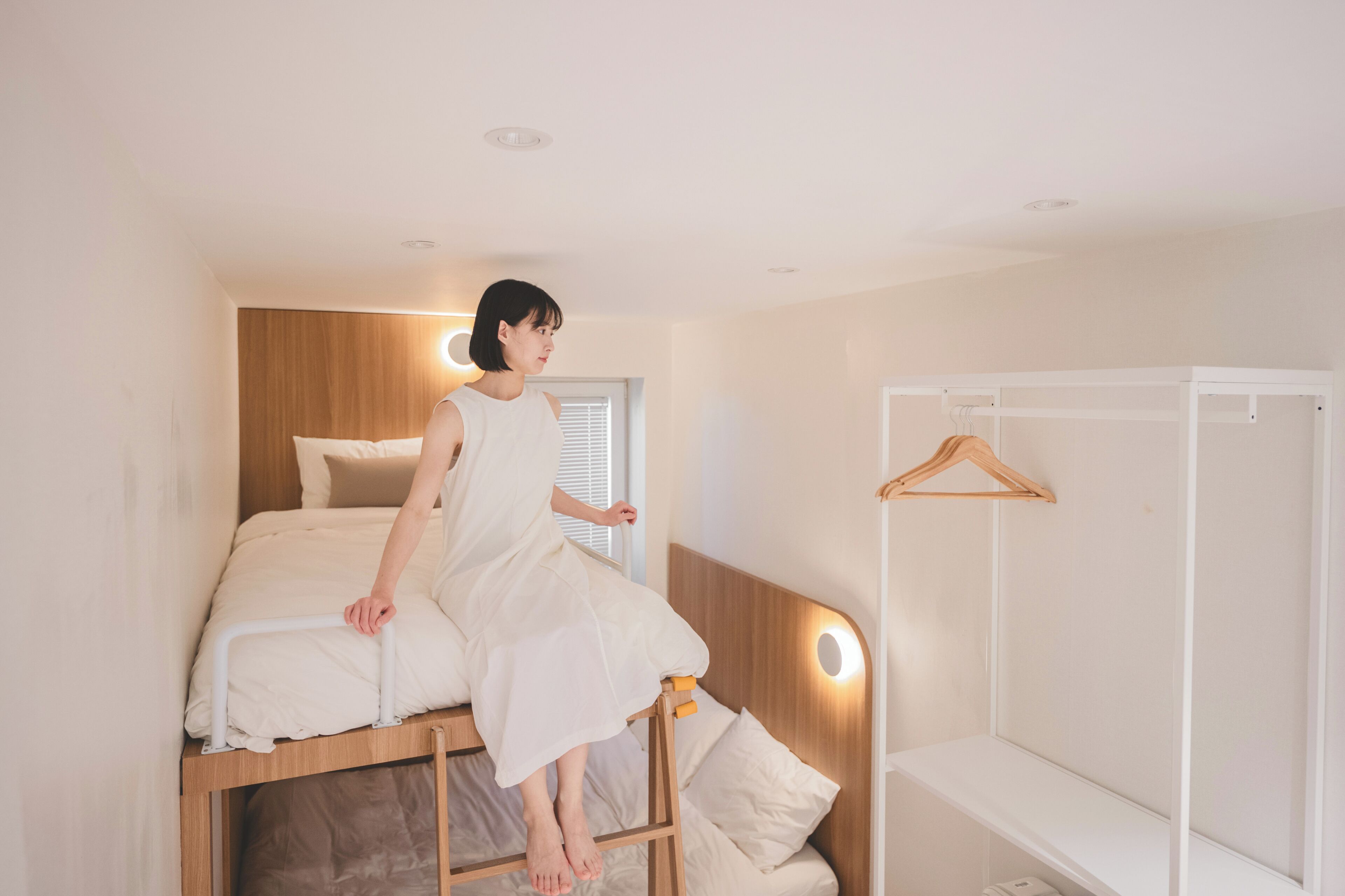 Una persona sentada en una escalera en un dormitorio con cabecera de madera, cama blanca y armario blanco.