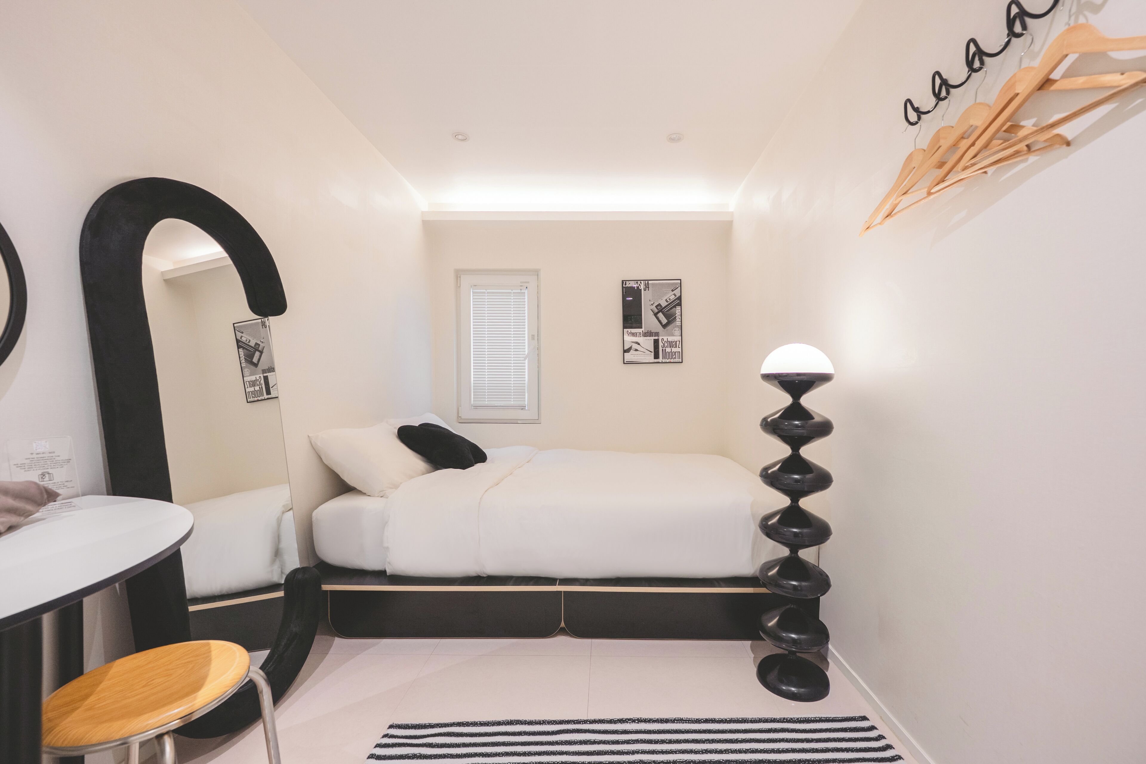 A bedroom with a bed, a desk, a chair, a mirror, a lamp, and a window.
