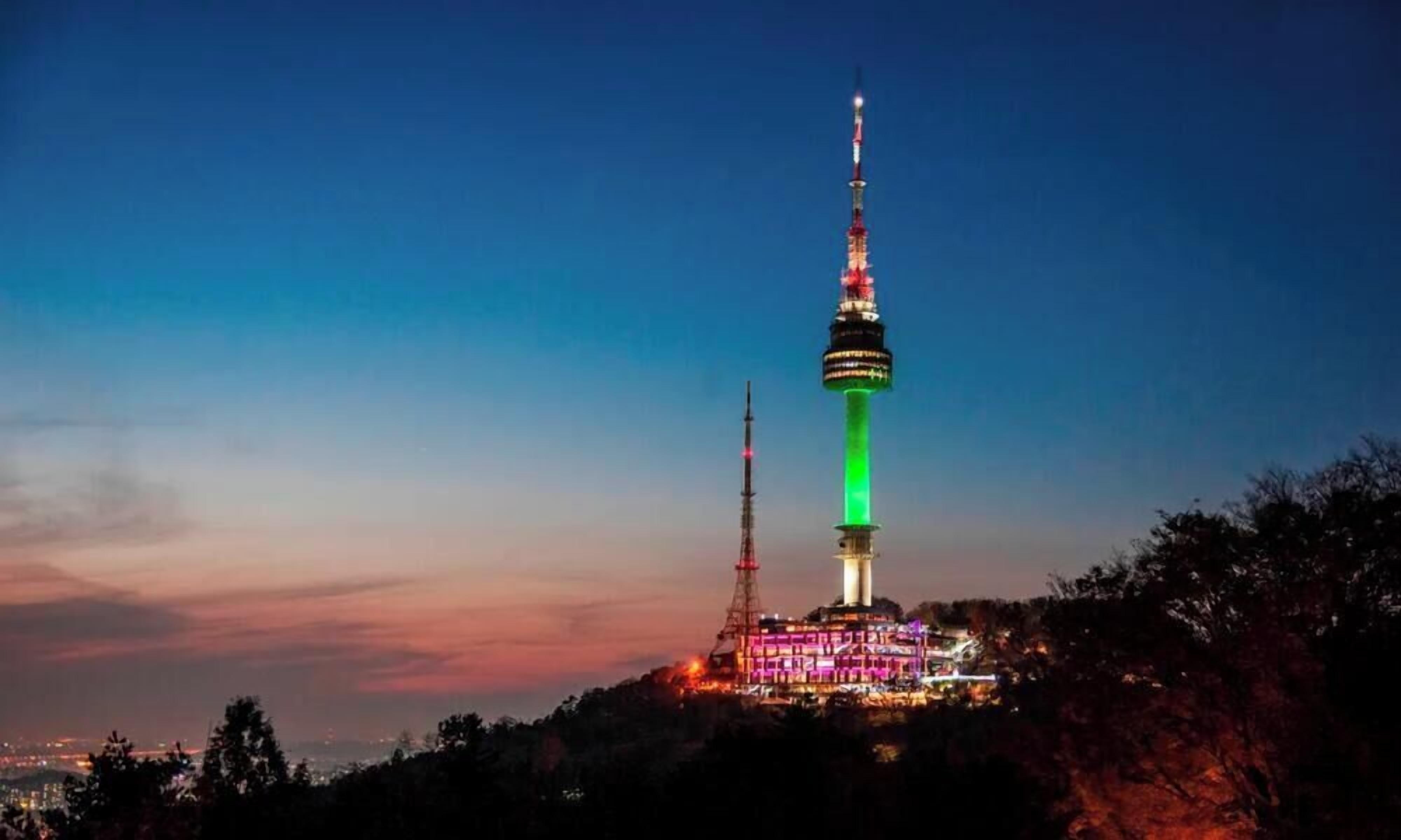 A tower with a green light at the base, surrounded by trees and buildings.