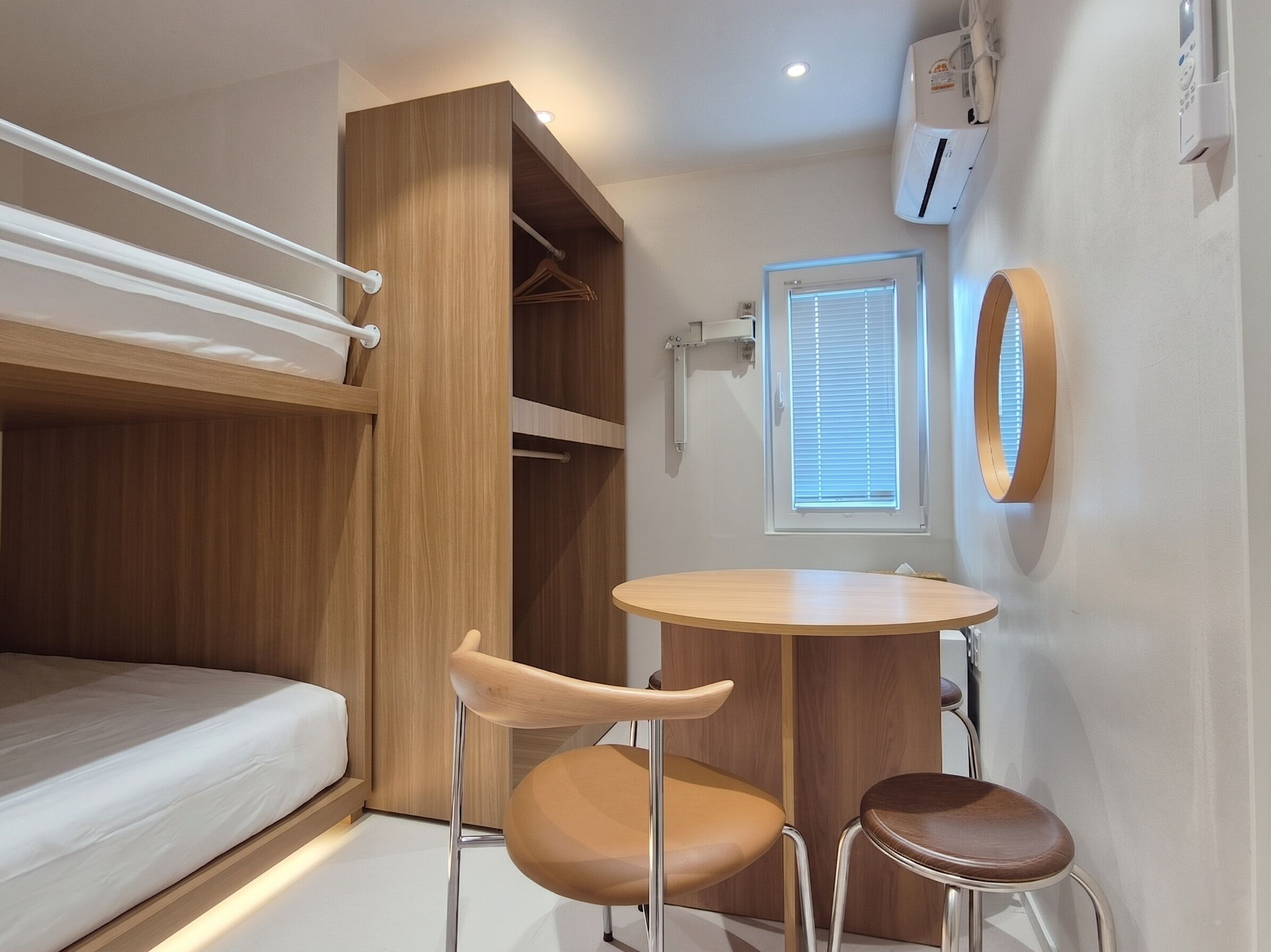 A compact room with a bunk bed, a round table with two chairs, a small window with blinds, and a wall-mounted air conditioning unit.