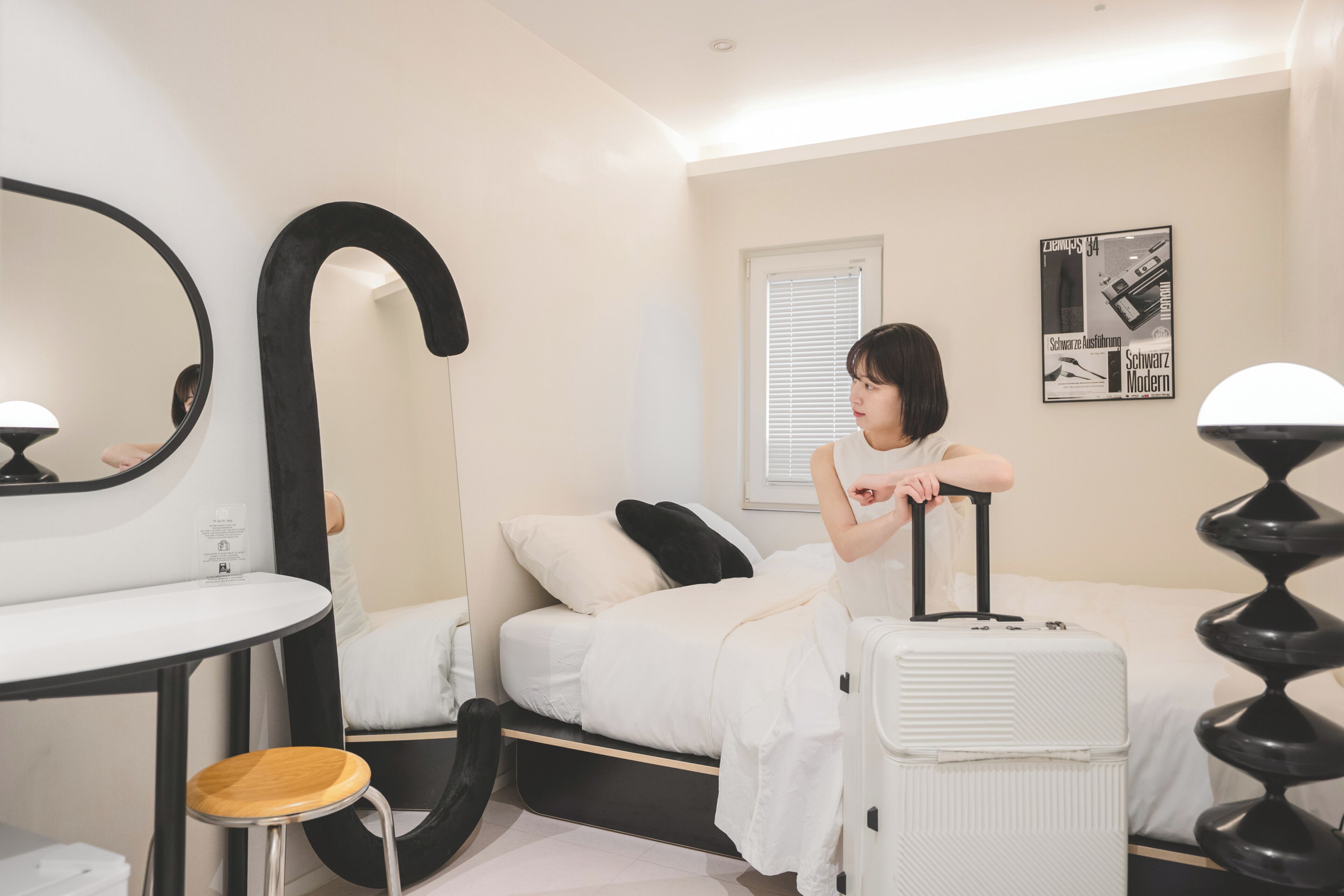 Een moderne hotelkamer met een bed, een ronde spiegel, een klein krukje, een witte koffer en een zwarte lamp.