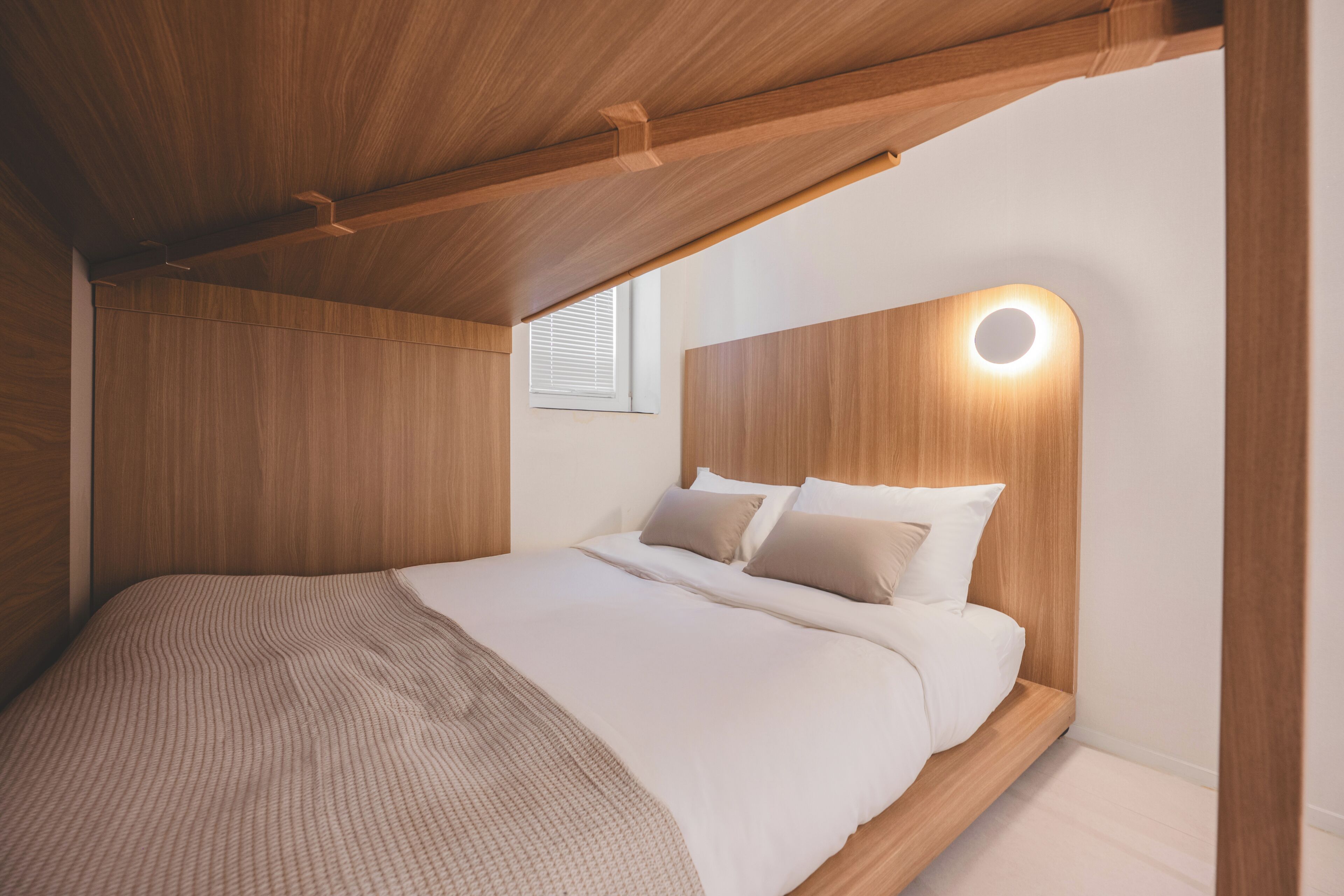 Une chambre avec un plafond en bois, un lit recouvert de draps blancs et un luminaire circulaire fixé au mur.