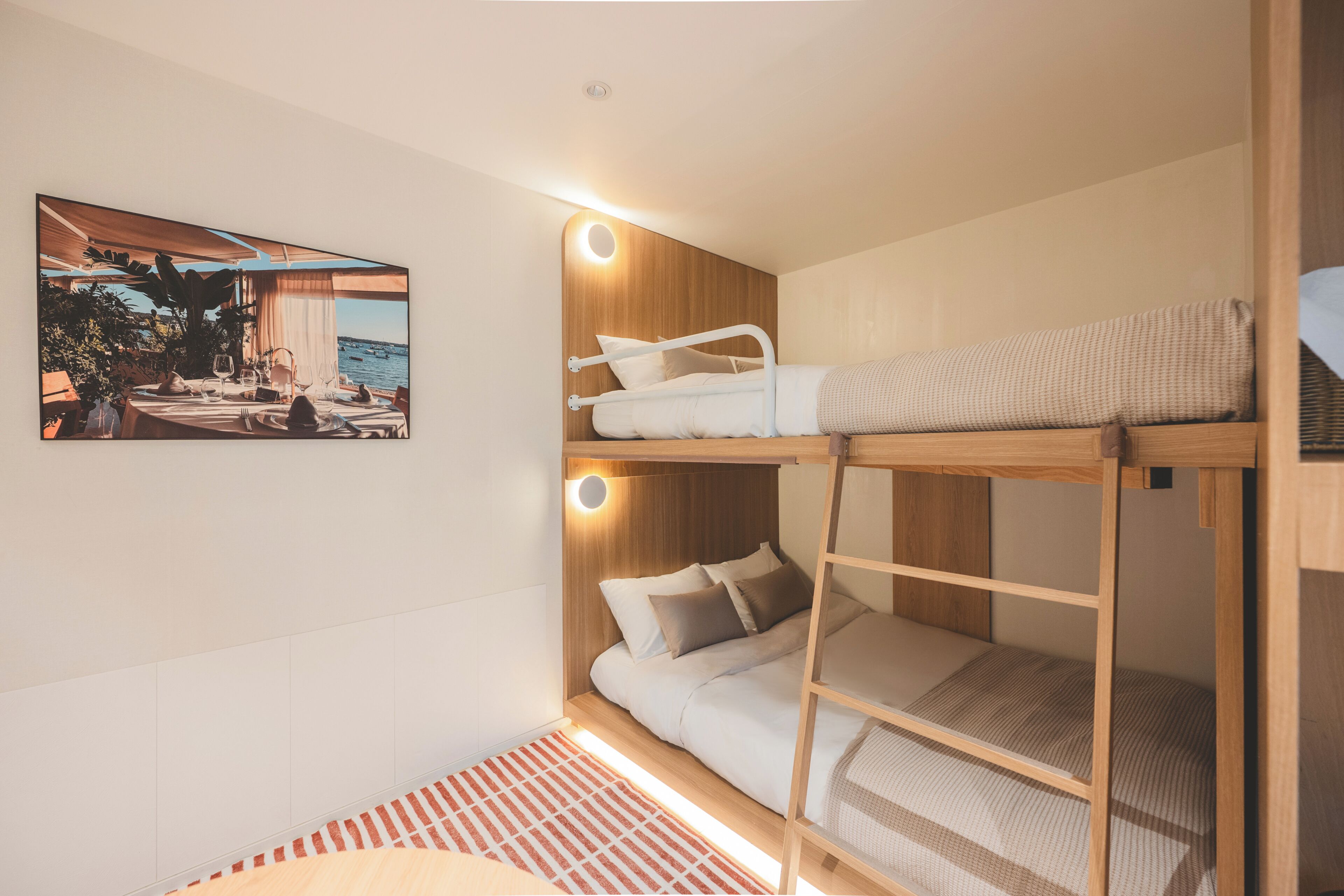 A bunk bed room with a view of the sea, a framed picture on the wall, and a small rug.