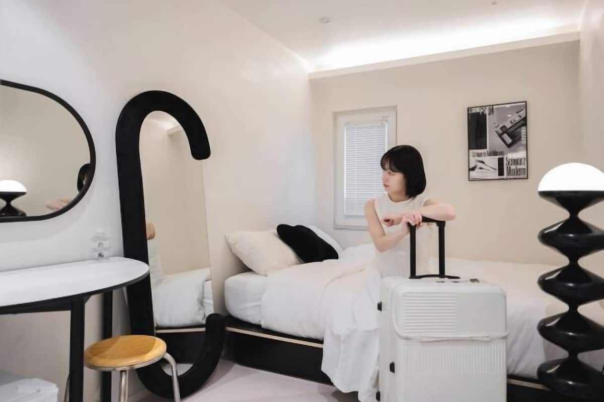 Un dormitorio con cama, mesita de noche, espejo, ventana y lámpara.