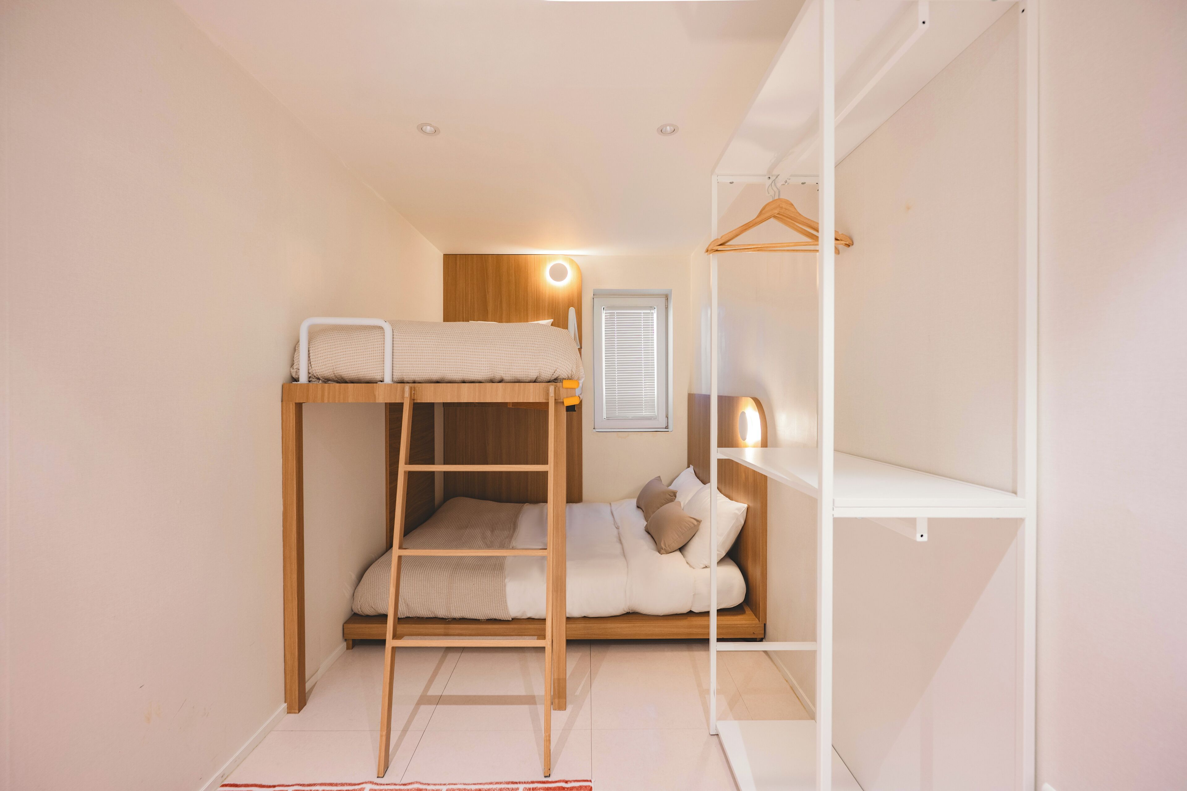 A bunk bed with a ladder, a bedside table, a hanging light, and a window with blinds.