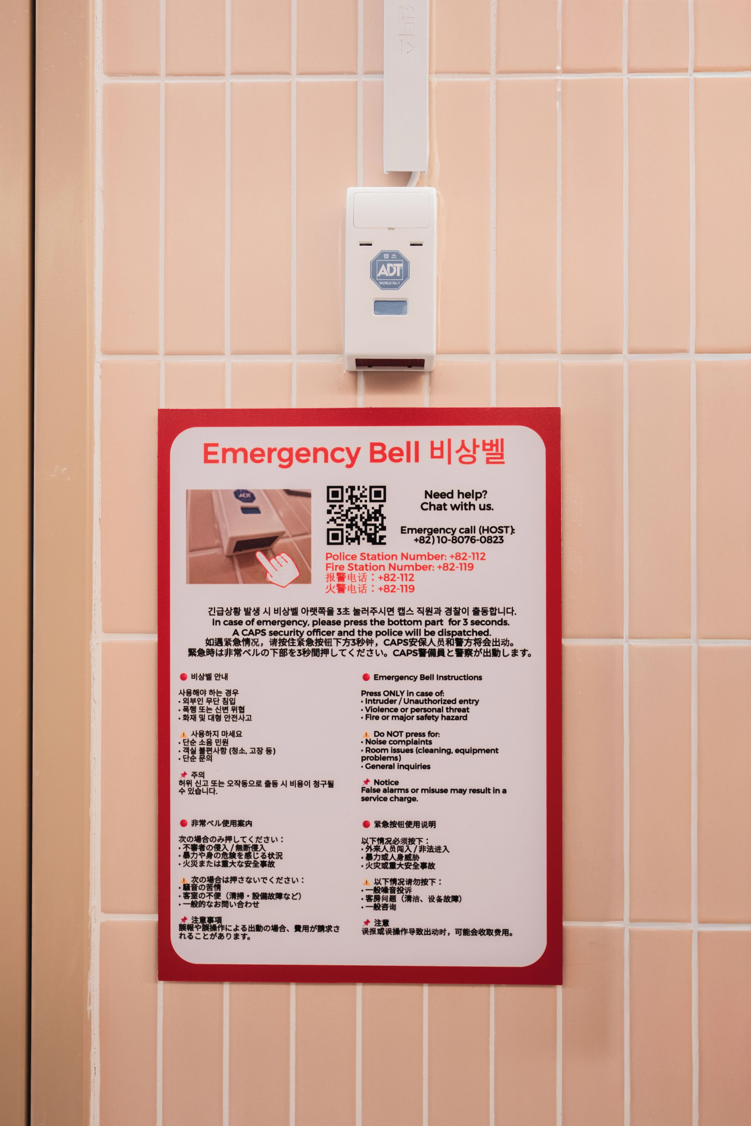 A wall-mounted emergency bell with instructions in multiple languages, a QR code, and contact numbers for police and fire stations.