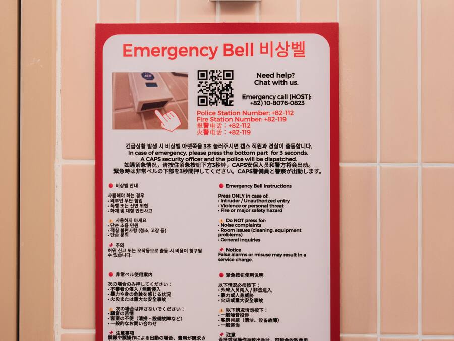 A wall-mounted emergency bell with instructions in multiple languages, a QR code, and contact numbers for police and fire stations.