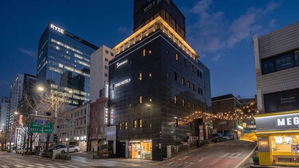 A modern hotel with a dark facade, multiple floors, and a prominent 'RYSE' sign on a neighboring building. - Slowon Hongdae (Seoul)