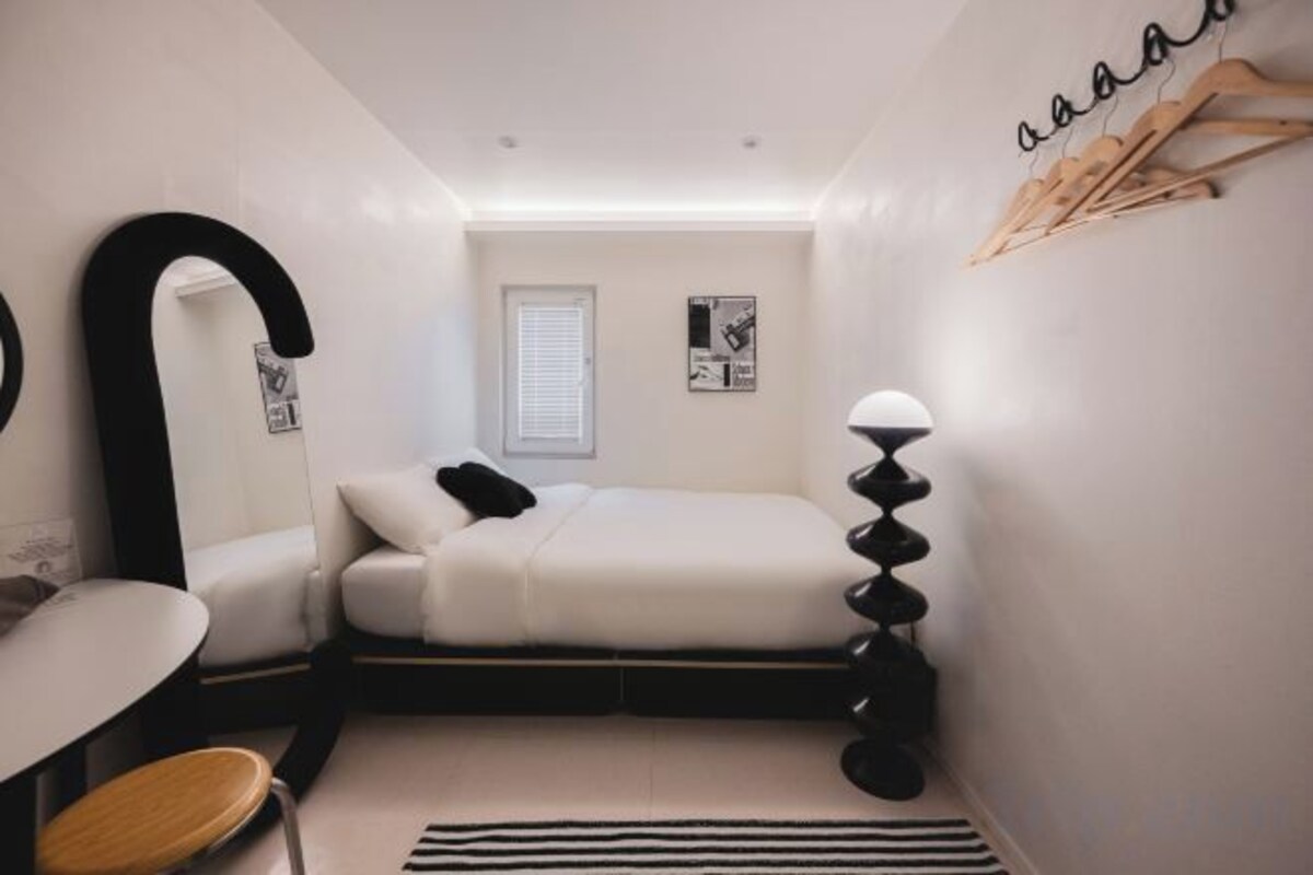 A bedroom with a bed, a chair, a desk, a mirror, and a window with blinds.