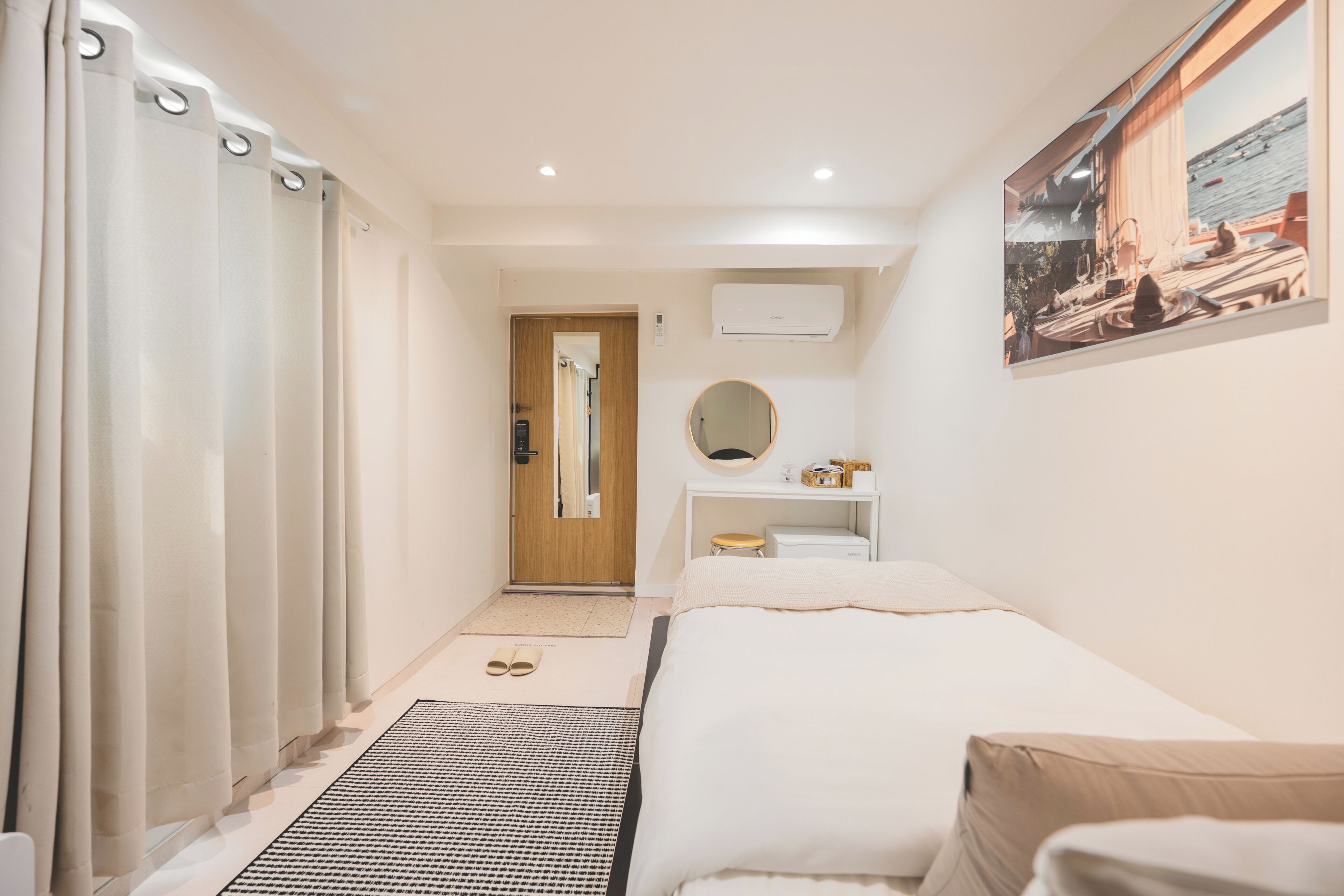 A hotel room with a bed, a desk with a mirror, a small stool, a window with a view of the sea, and a door leading to another room.