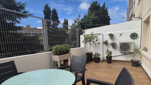 A rooftop patio with a table and chairs, potted plants, and a white wall with an air conditioning unit.