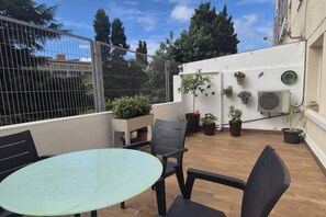 Une terrasse sur le toit avec une table et des chaises, des plantes en pot et un mur blanc sur lequel est installé un appareil de climatisation.