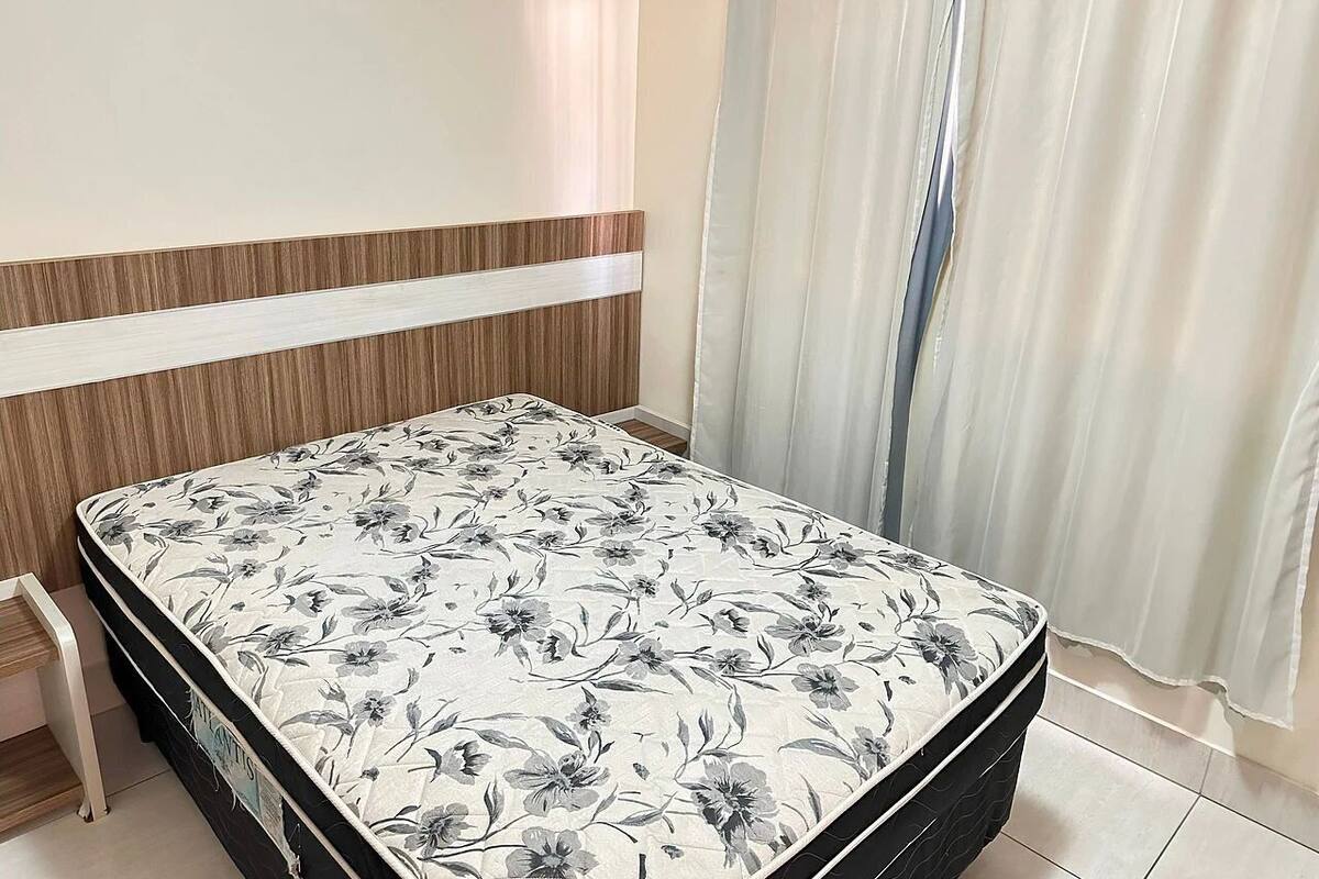 Un llit amb llençols florals en una habitació amb un capçal de fusta, una taula petita i una finestra amb cortines.