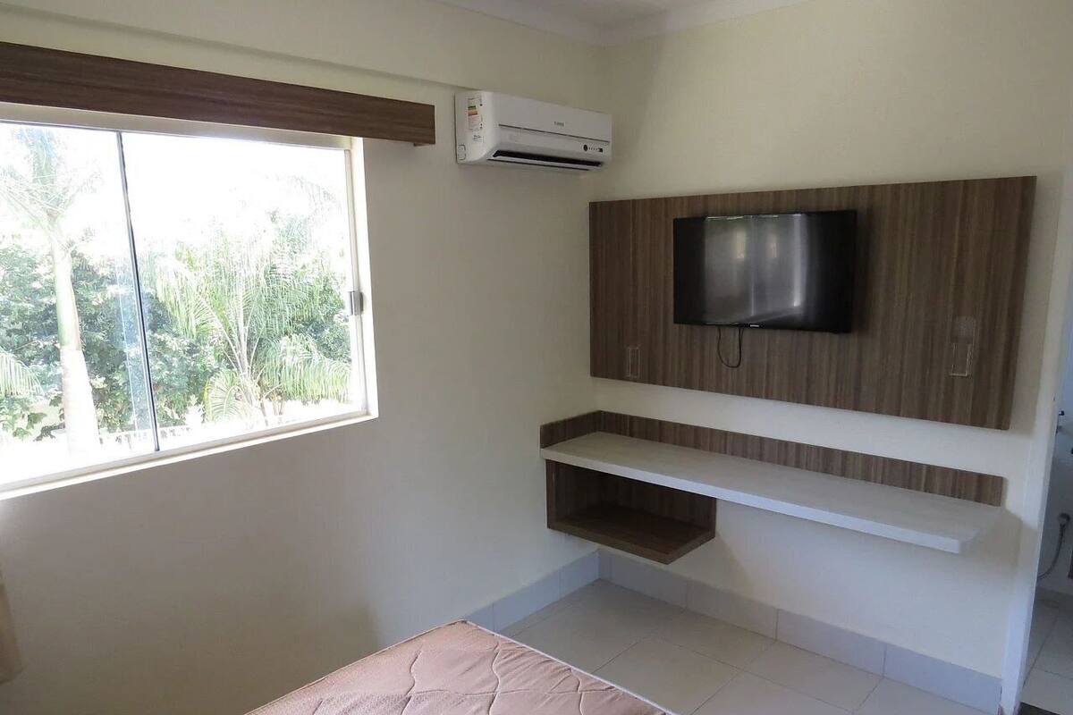 Habitació amb un llit, un televisor fixat a un panell de fusta, una estant i un aparell d'aire condicionat.