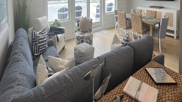 A spacious living room with a large gray sectional sofa, a dining area with a wooden table and chairs, and a view of the marina through large windows. - Brand new Bayfront Luxury Townhome with Pool and Elevator. (Spray Beach)
