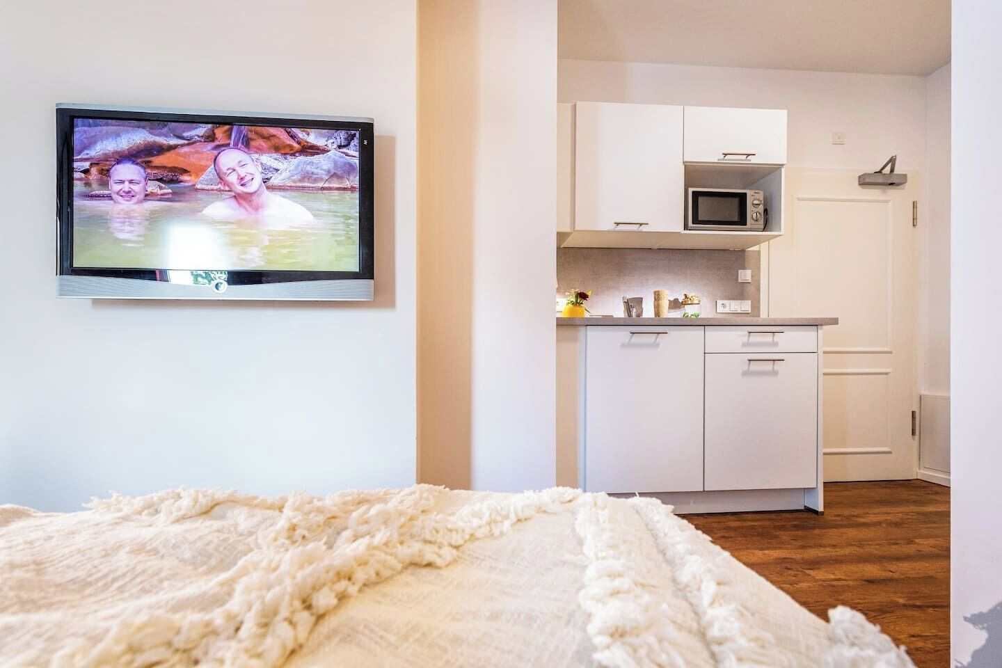 Een slaapkamer met een bed, een televisie aan de muur en een kitchenette met kastjes en een magnetron.