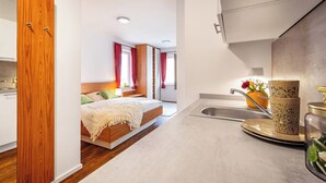 A modern hotel room with a wooden wardrobe, a bed with a white bedspread, a small kitchenette with a sink and a vase of flowers.