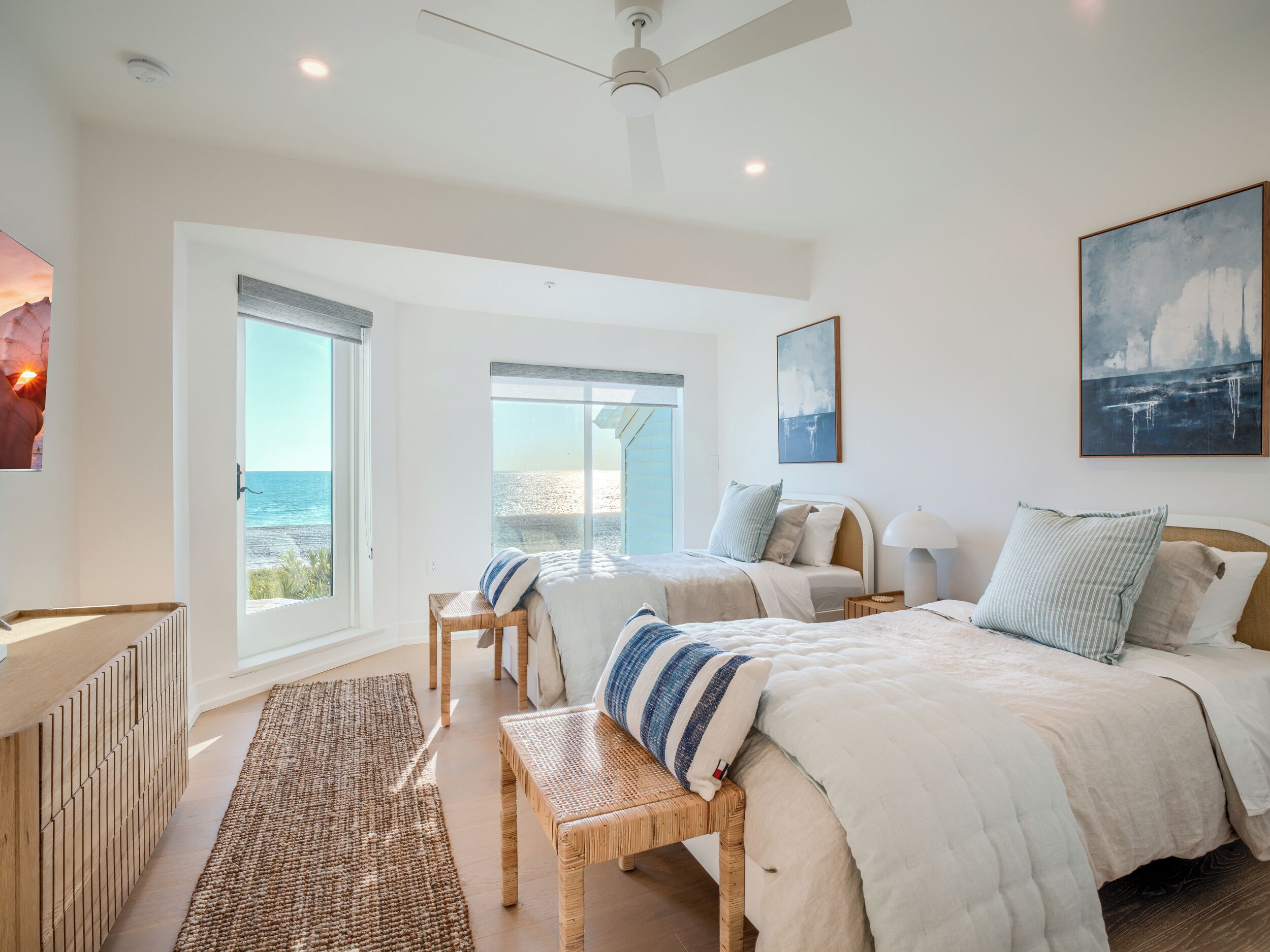 A bedroom with a large window overlooking a beach, a ceiling fan, and a bed with multiple pillows.