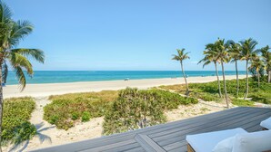 Eine Holzterrasse mit einem weißen Sofa und Kissen, mit Blick auf einen Strand mit Palmen und wolkenlosem, blauem Himmel.