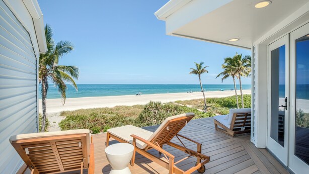 Eine Holzterrasse mit einem weißen Tisch und zwei Holzsesseln, mit Blick auf einen Strand mit Palmen und dem Meer.