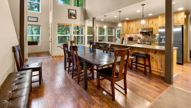 A spacious dining area with a long wooden table and chairs, a kitchen with wooden cabinets and stainless steel appliances, and large windows with blinds. - Evergreen House in heart Government Camp (Government Camp)