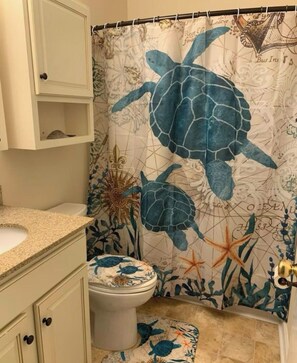 Un baño con cortina de ducha con forma de tortuga marina, un inodoro con tapa decorativa y un pequeño estante sobre el lavabo.
